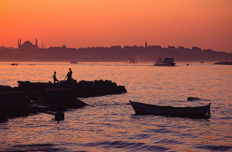 Two children silhouetted on rocks at a crimson Bosphorus sunset with Blue Mosque and Hagia Sophia on the Istanbul skyline, a wooden rowing boat in the foreground, fine art photography print