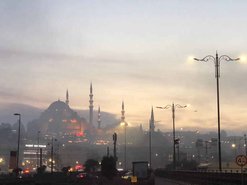 Süleymaniye Mosque dome and minarets glowing amber through thick morning mist at dawn over Istanbul, with street lights still burning on Galata Bridge below, fine art photography print