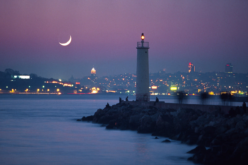 Crescent moon in a violet night sky above an Istanbul lighthouse on the breakwater, with Galata Tower glowing and the lit European shore of Istanbul reflected on the Bosphorus, fine art photography print