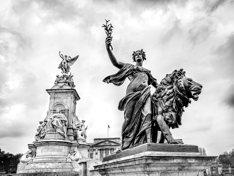 Low angle black and white fine art print of the Victoria Memorial bronze Progress figure raising a laurel branch with a lion beside her, Queen Victoria enthroned behind and Buckingham Palace in the background under dramatic clouds